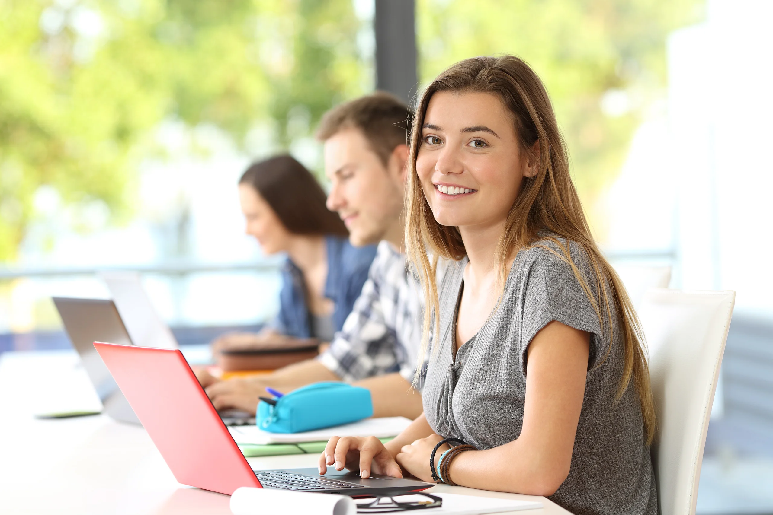 Students working at laptops in a well-lit classroom. The focal point is a happy girl looking at the camera.