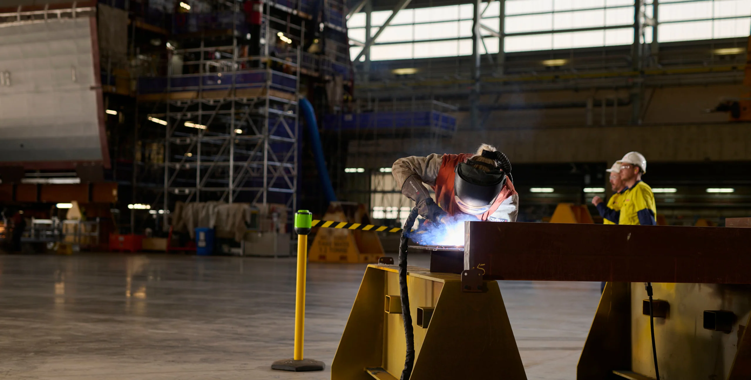 Welder working at shipyard. A large, incomplete hull is visible in the background.