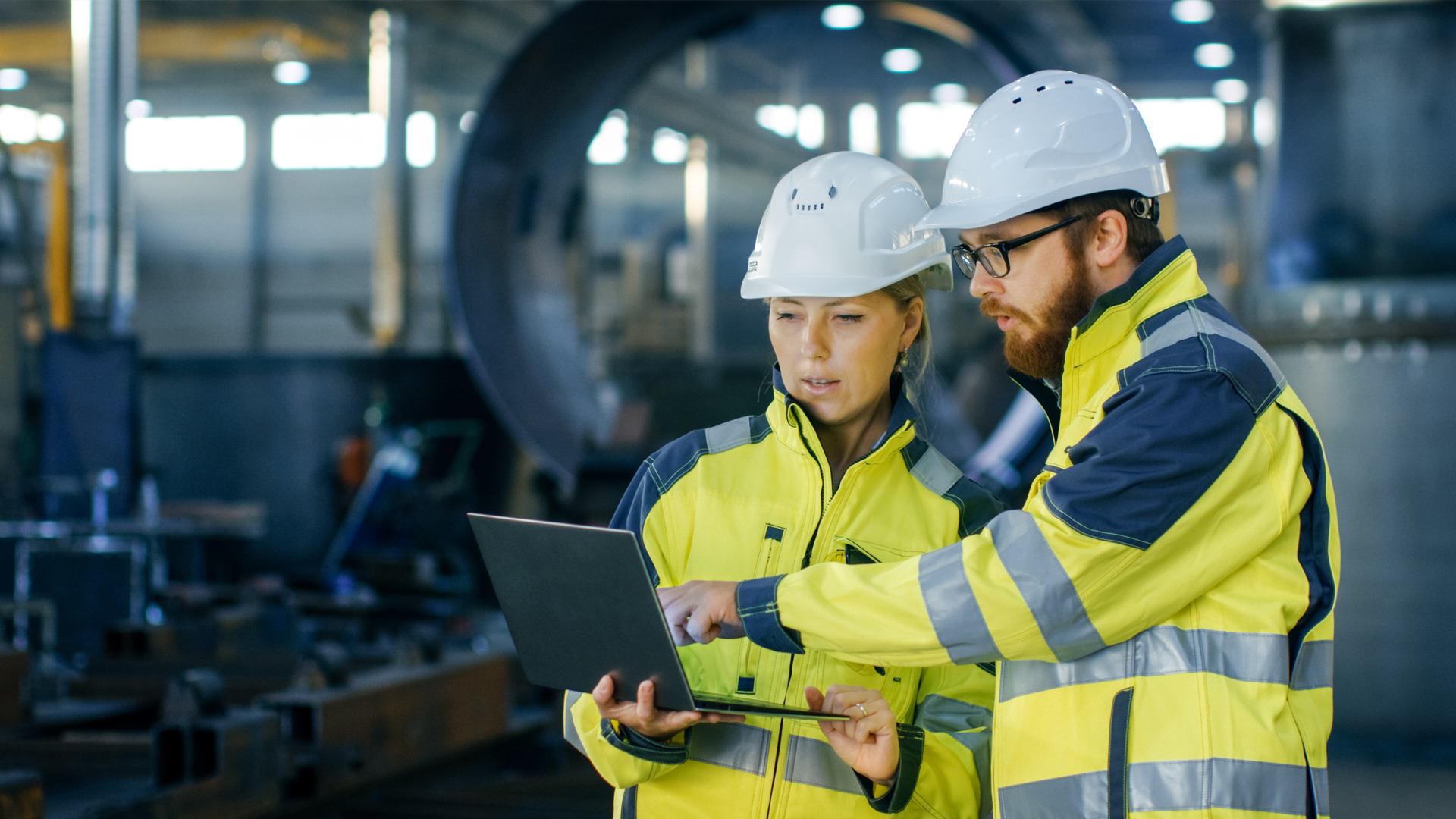 Man and woman wearing high‑visibility clothing and hard hats in a warehouse, standing together and looking at a screen.