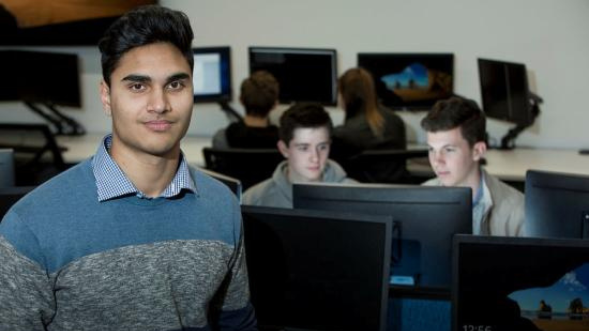 Australian Industry Capability (AIC) - Foreground man looks at camera. Background two men sit at a computer working cooperatively