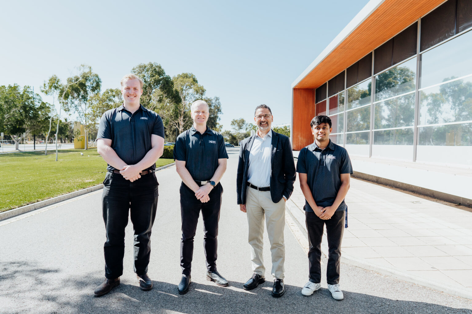 Four men stand outside on a sunny day and smile for the camera.