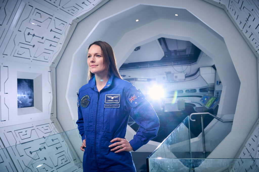 Katherine Bennell-Pegg stands in a space craft hallway in her iconic blue space jumpsuit